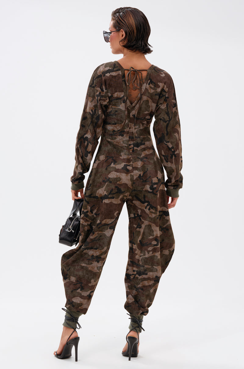 A person faces away, wearing the COMFY COZY SWEATSHIRT JUMPSUIT IN CAMO with black high-heeled sandals and sunglasses, holding a small black handbag against a plain white background.