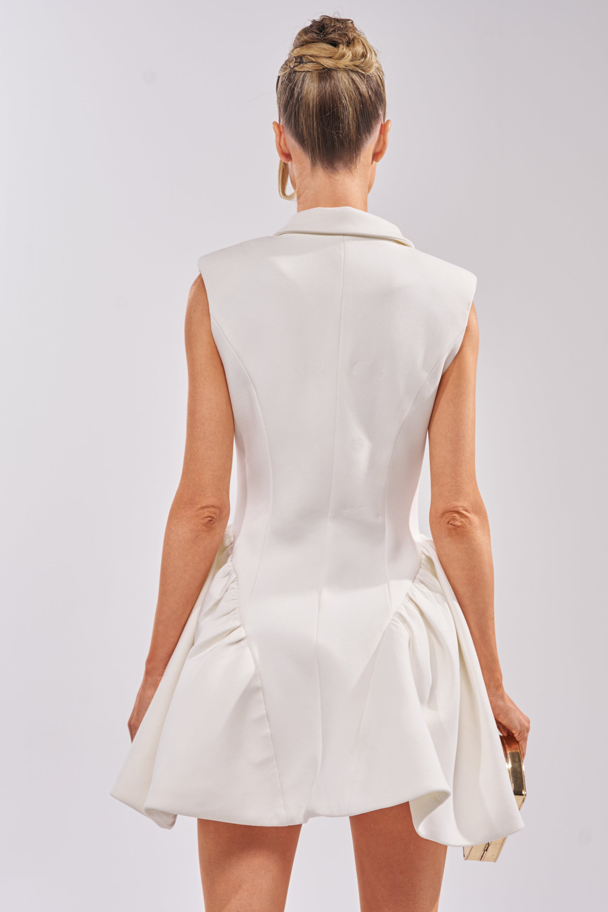 A woman with blonde hair in an elegant updo is seen from the back against a plain light background, wearing the REPRESENTING ME SLEEVELESS BLAZER DRESS IN WHITE and holding a small rectangular clutch in her right hand. 4