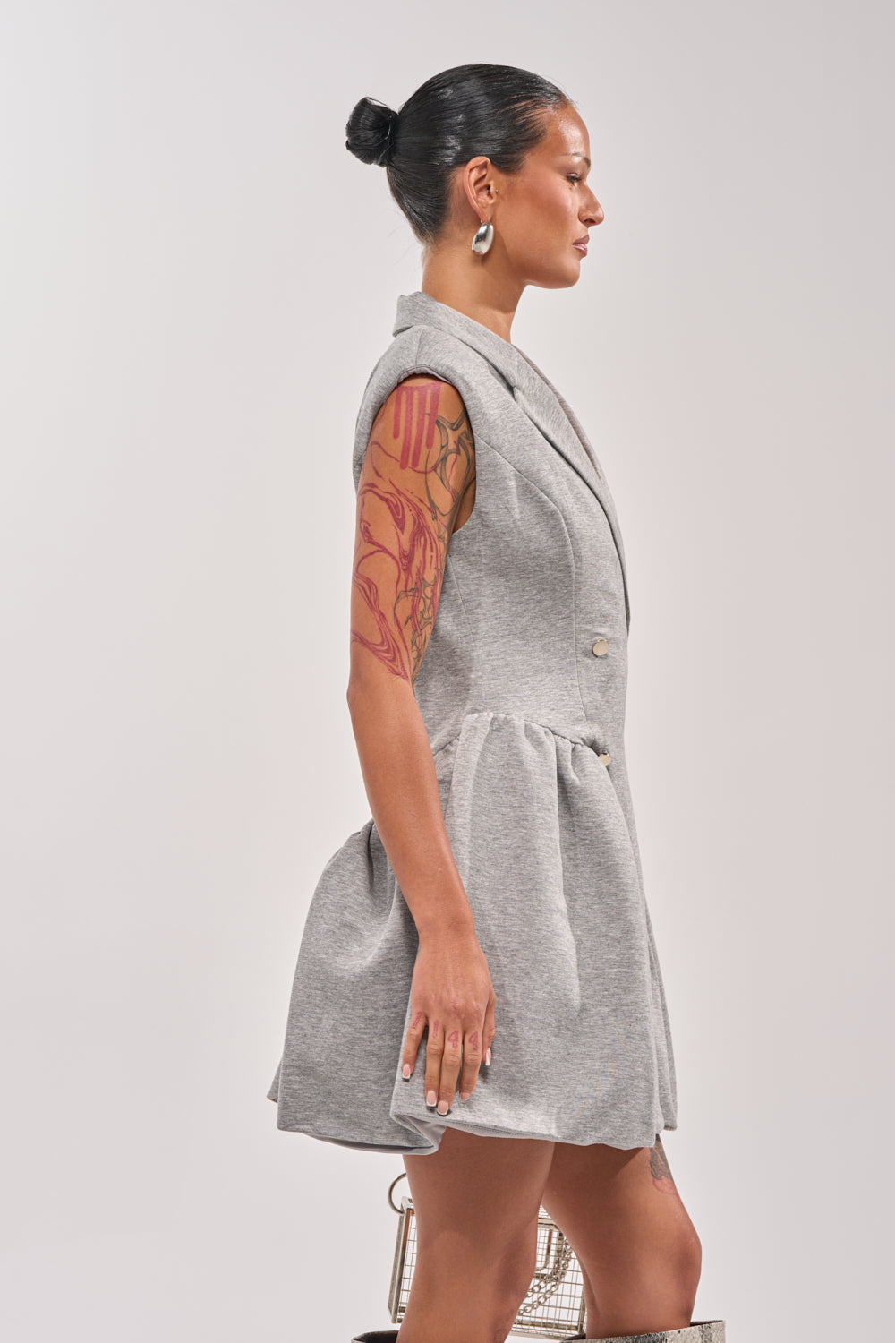 A woman with a tattooed arm and hair in a bun wears the IT SUITS YOU SLEEVELESS BLAZER MINI DRESS, a chic grey fit-and-flare sleeveless blazer dress with a flared skirt. She stands in profile against a plain, light background. 3