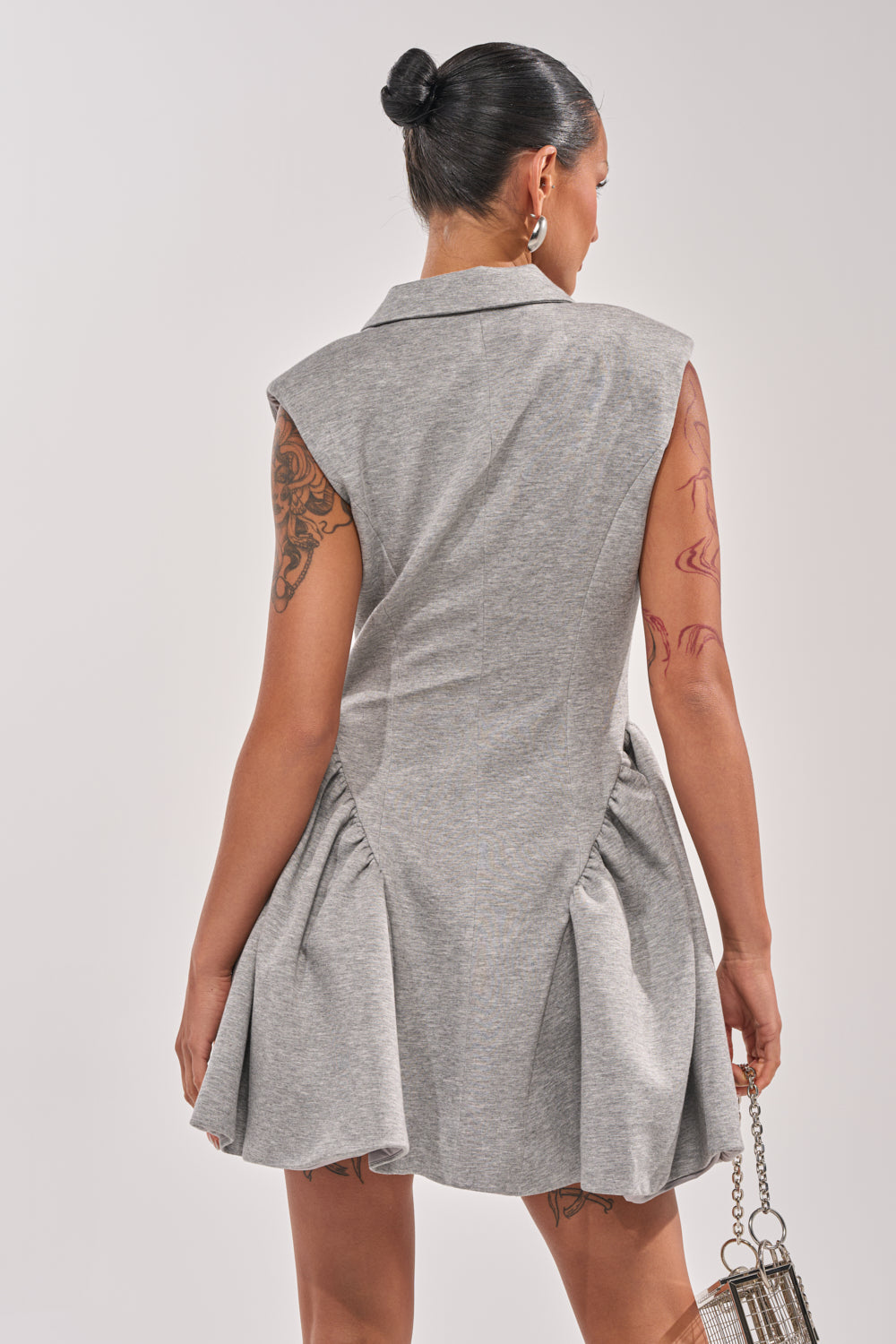 A woman with dark hair in a bun is seen from behind, wearing the IT SUITS YOU SLEEVELESS BLAZER MINI DRESS in grey with a flared skirt. She has tattoos on her arms and carries a small metallic handbag. 4
