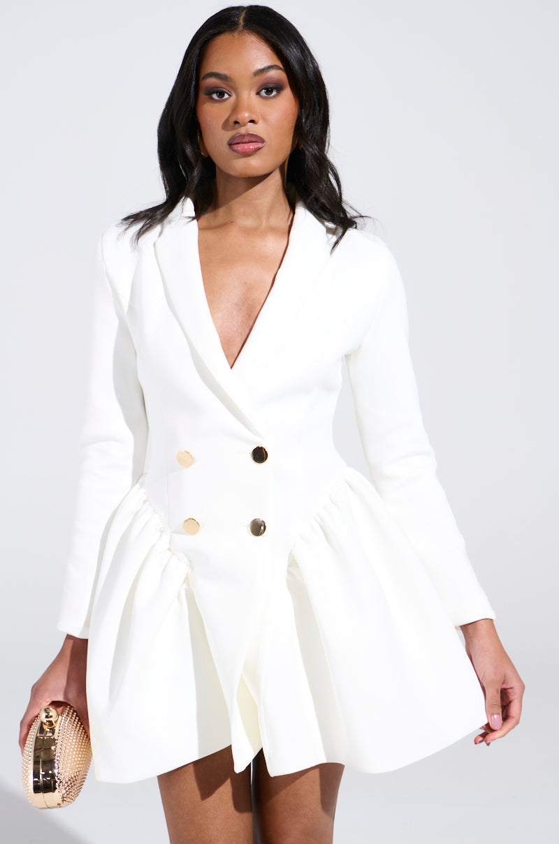 Wearing the HBIC BLAZER MINI DRESS IN WHITE with gold buttons, she holds a metallic gold clutch. Her long, wavy dark hair flows over the scuba fabric as she poses against a plain light background.