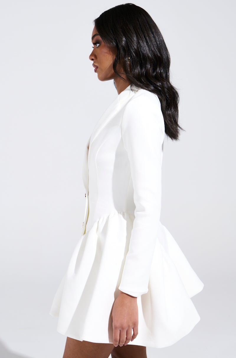 A woman with long, wavy black hair is shown in profile wearing the HBIC BLAZER MINI DRESS IN WHITE, featuring a chic blazer style and flared pleated skirt crafted from sleek scuba material, against a plain light background.
