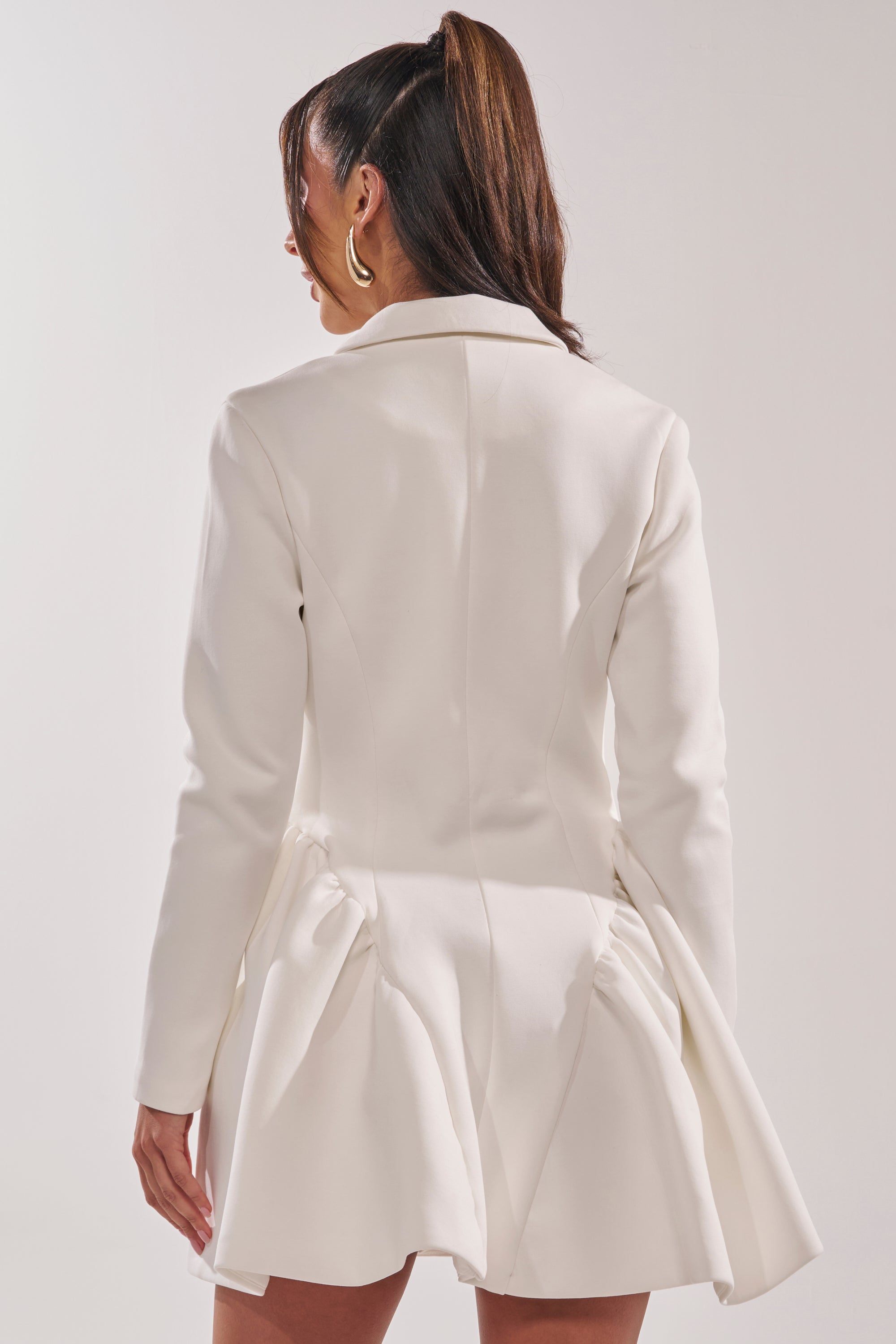 A woman faces away, highlighting the back of the HBIC Blazer Mini Dress in White featuring a pleated skirt. Her high ponytail and large hoop earrings give this tailored blazer dress a chic, modern vibe. 3