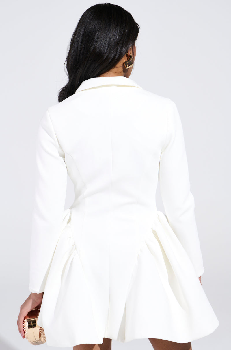 A woman with long dark hair, seen from behind, wears the HBIC BLAZER MINI DRESS IN WHITE—a chic mini dress with long sleeves and a flared skirt. She holds a small gold clutch and accessorizes with gold hoop earrings.