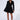 A model stands against a plain background wearing the HBIC Blazer Mini Dress, featuring long sleeves, button details, and a flared skirt. She has curly hair and is looking down. She pairs the dress with white socks and black strappy high-heeled sandals.