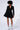 A model stands against a plain background wearing the HBIC Blazer Mini Dress, featuring long sleeves, button details, and a flared skirt. She has curly hair and is looking down. She pairs the dress with white socks and black strappy high-heeled sandals.