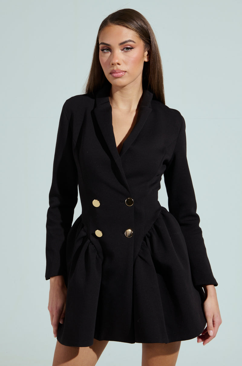 A woman stands against a light background in an HBIC BLAZER MINI DRESS, featuring gold buttons and a black, double-breasted design. Her long, straight hair complements her neutral expression, while the peplum hem adds flair to the dress's flared skirt starting at the waist.