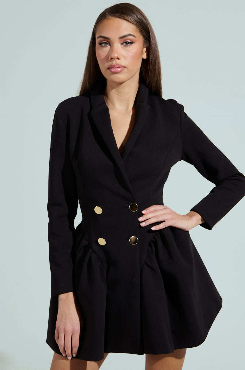 A woman confidently wears the HBIC BLAZER MINI DRESS, a chic black design with double-breasted gold buttons. Her long, straight hair cascades down as her neutral expression exudes poise against a soft, light background.