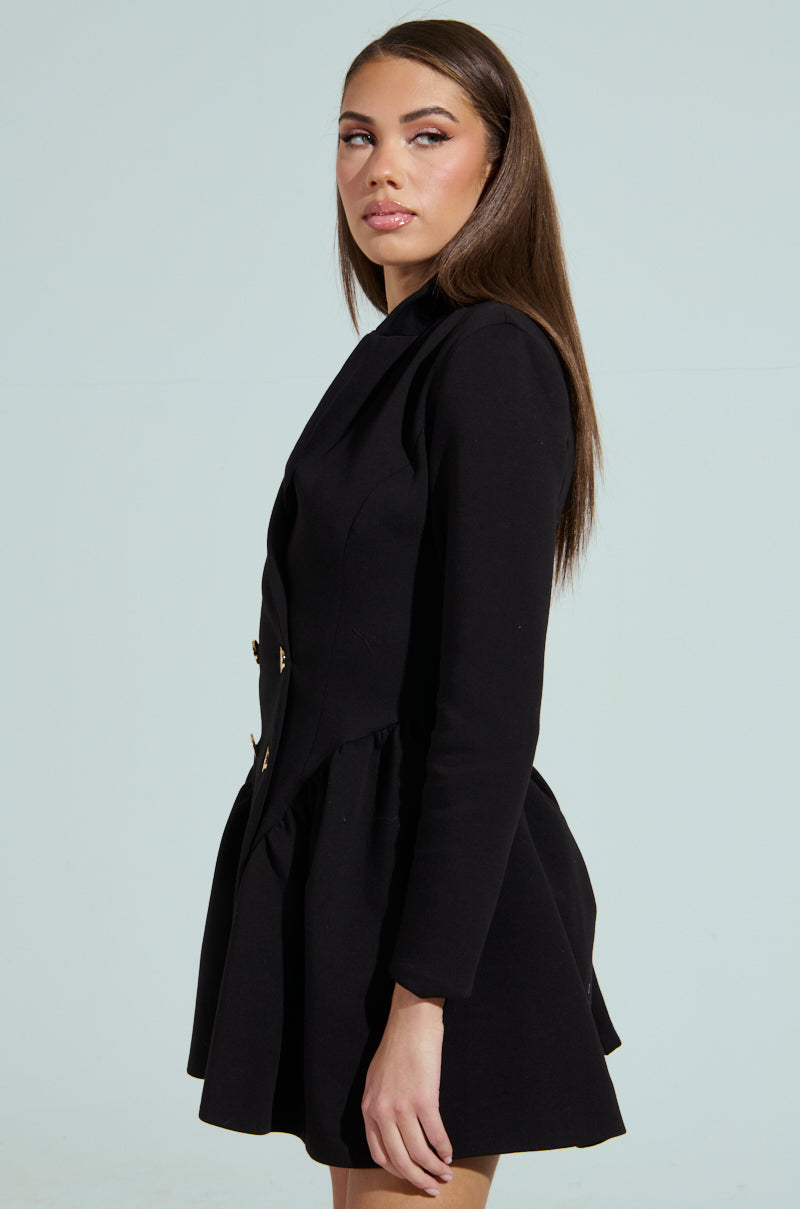 A woman with long brown hair stands in profile against a light gray background, wearing the HBIC BLAZER MINI DRESS—an elegant black double-breasted blazer mini dress with buttons. Her gaze over her shoulder is calm and composed.
