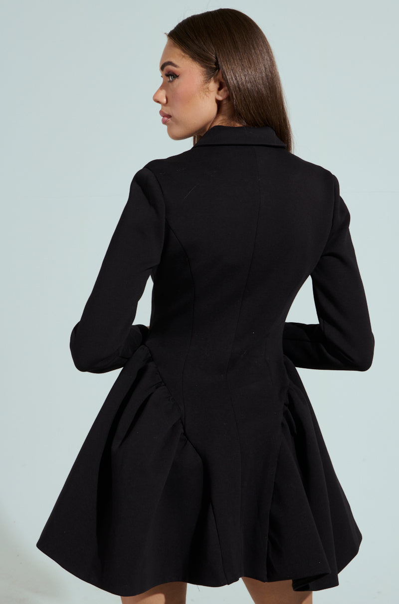 A woman wears the HBIC BLAZER MINI DRESS, a fitted black coat dress with long sleeves and a peplum hem, while looking to the side against a plain light background.