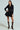 A woman poses in the HBIC BLAZER MINI DRESS, featuring a black double-breasted design with gold buttons and a pleated skirt. She's wearing sheer black knee-high socks and black heels with gold tips, standing against a plain light green background with her hand on her hip.