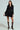 Dressed in the HBIC Blazer Mini Dress with gold buttons, a woman pairs it with sheer knee-high socks and black heels featuring gold toe caps. Her stylish look is emphasized as she poses elegantly against a light gray background, exuding sophistication.