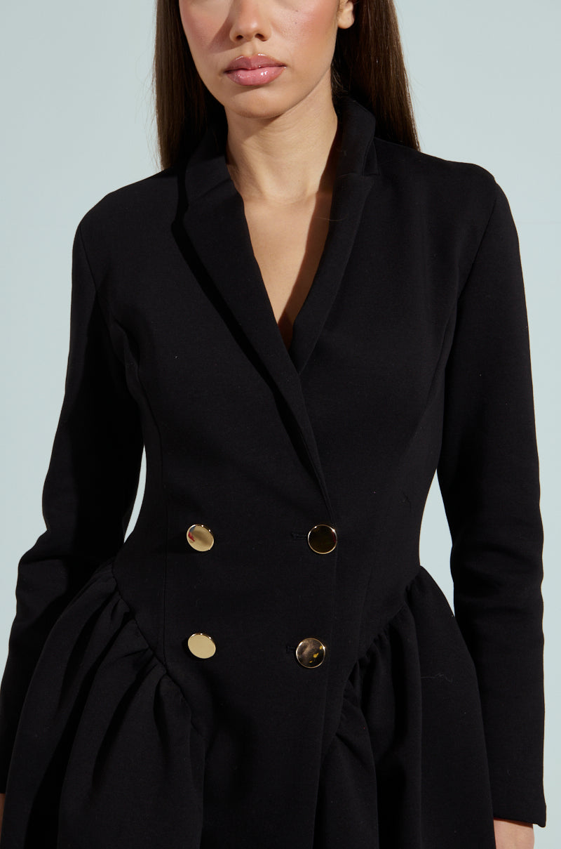 A woman in the HBIC BLAZER MINI DRESS, a black double-breasted coat with gold buttons and structured fabrication, featuring ruched waist detailing against a light gray background.