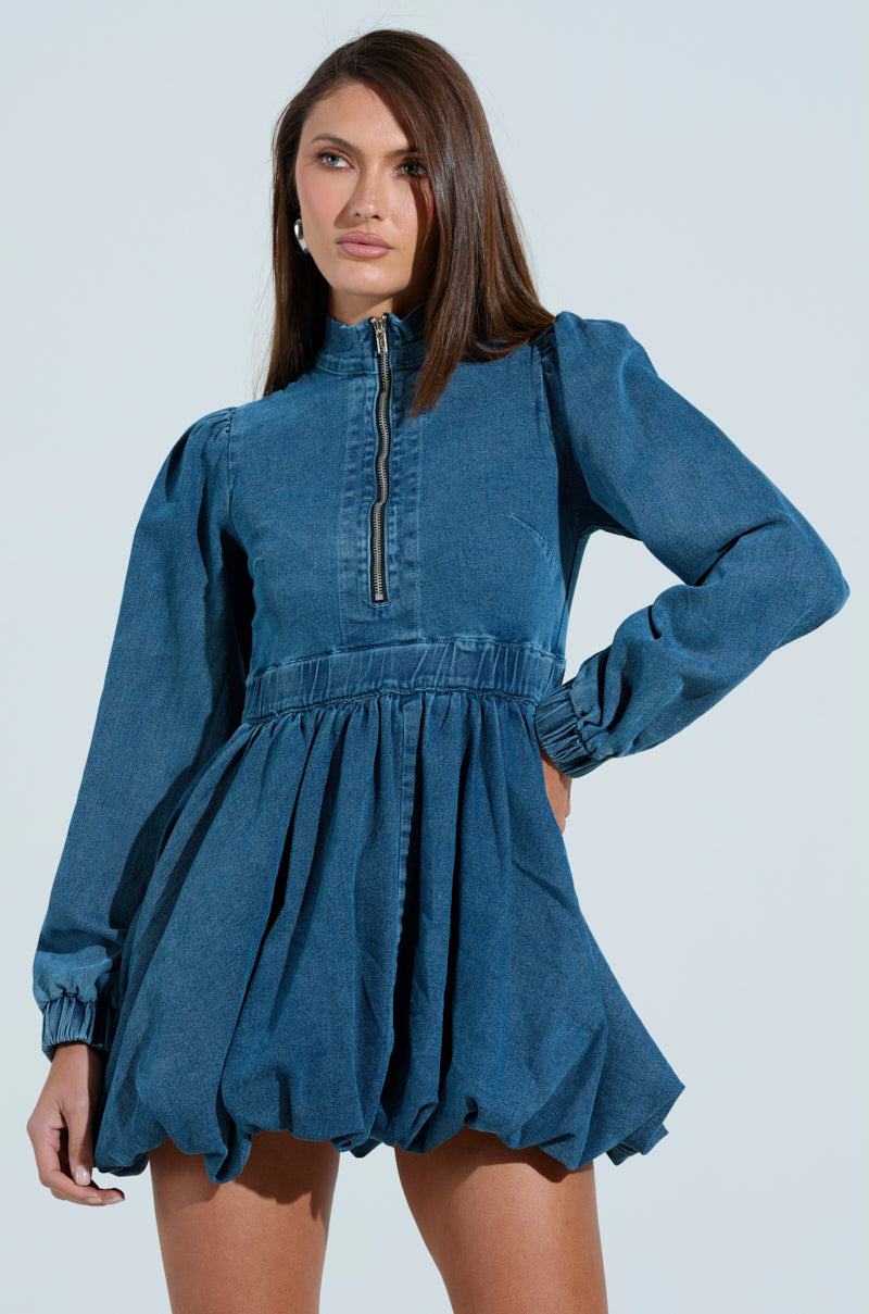 In a PASS THE BUBBLY denim mini dress featuring long sleeves, an adjustable zipper closure, and a gathered waist, a woman poses confidently against a plain background. Her straight hair and pearl earrings add elegance to her chic look.