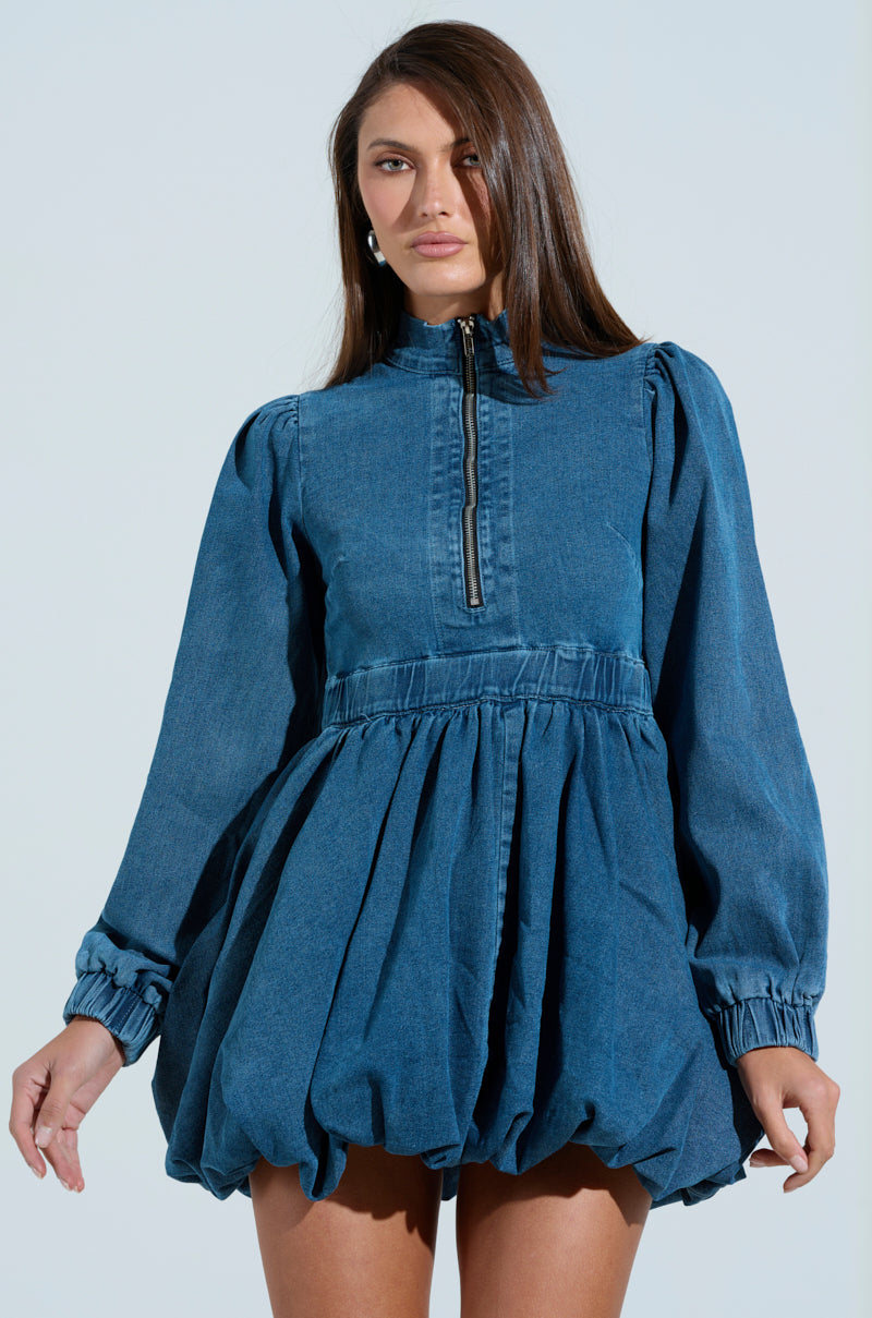 A person with long brown hair stands against a light backdrop, wearing the PASS THE BUBBLY DENIM MINI DRESS. This stylish mini dress features puffed long sleeves, an adjustable zipper closure, a gathered waist, and a bubble hem skirt. The person is looking forward.