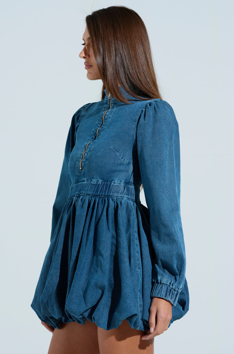 A woman wears the PASS THE BUBBLY DENIM MINI DRESS, showcasing a long-sleeved, high-neck design with a fitted bodice and skater silhouette. Her long, straight hair cascades as she looks left against a simple backdrop.