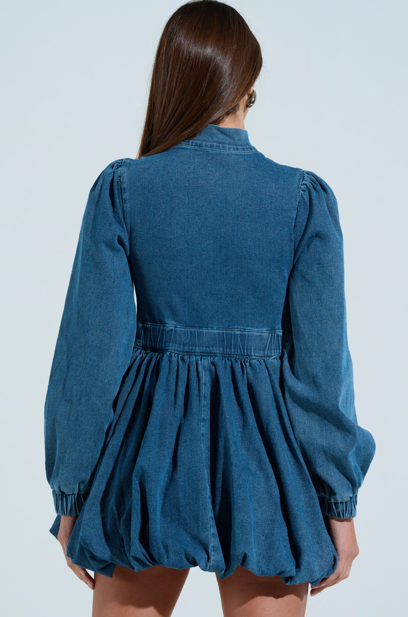 Against a light blue backdrop, a person with long hair presents their back to the camera, donning the PASS THE BUBBLY DENIM MINI DRESS. With puffy long sleeves and a gathered skirt, this skater dress silhouette enhances the breezy ambiance.