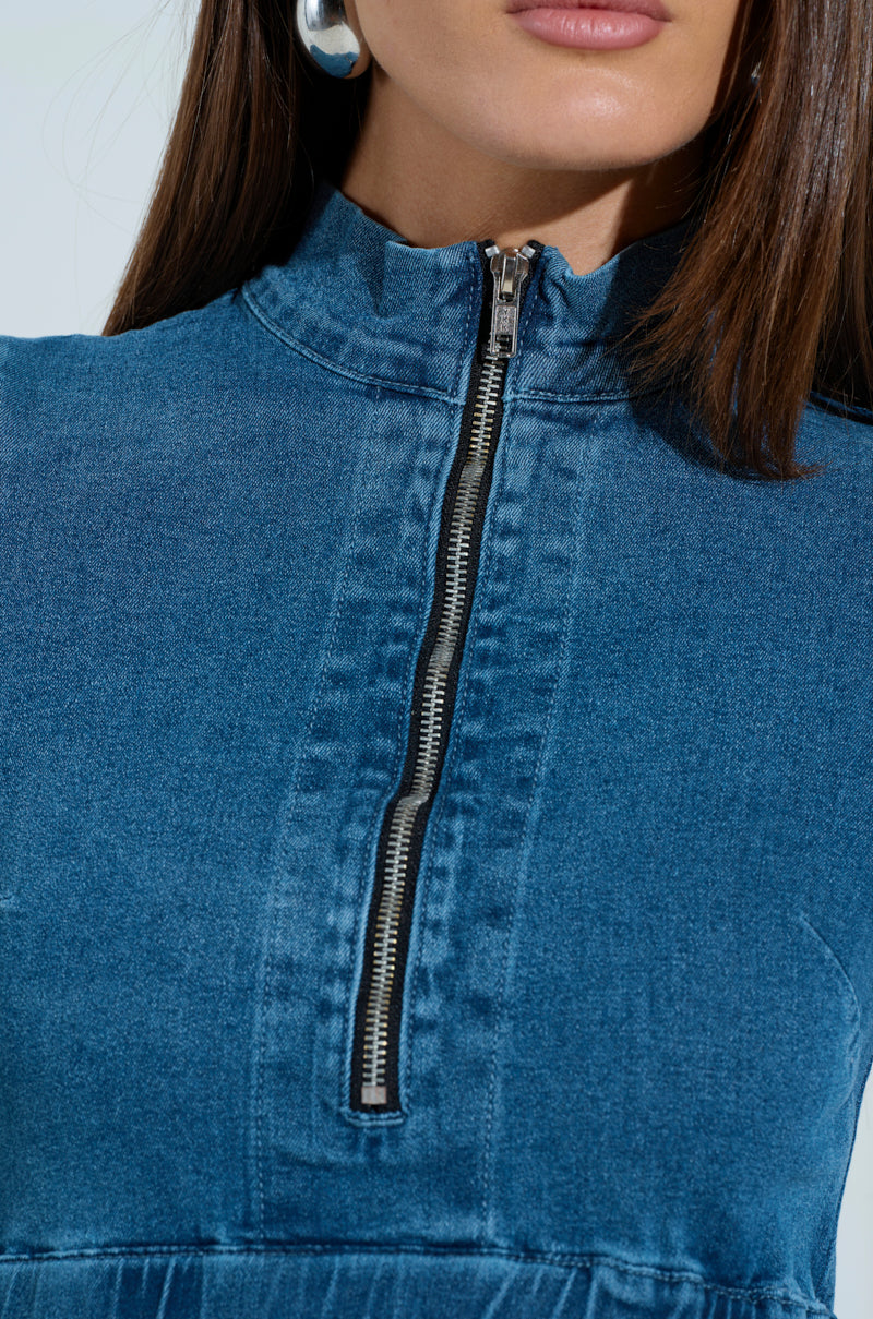 A close-up of someone wearing the "PASS THE BUBBLY" denim mini dress with a bubble hem and a center zipper. It features a high collar, complemented by silver hoop earrings. Their straight brown hair contrasts against the plain white background.