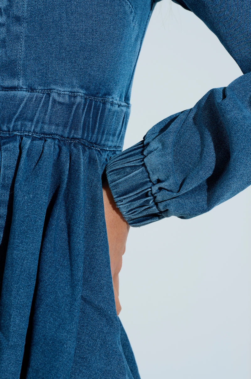 Close-up of a person in the PASS THE BUBBLY DENIM MINI DRESS with their hand on their hip. It has a gathered waist, long sleeves with elastic cuffs, and a chic bubble hem silhouette against a light blue background.