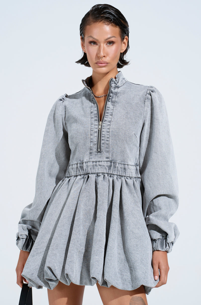 A woman in the PASS THE BUBBLY denim mini dress in light grey, featuring a zippered neckline and skater silhouette. Her hair is slicked back while holding a black handbag against a simple, light background that accentuates the dress's chic appeal.