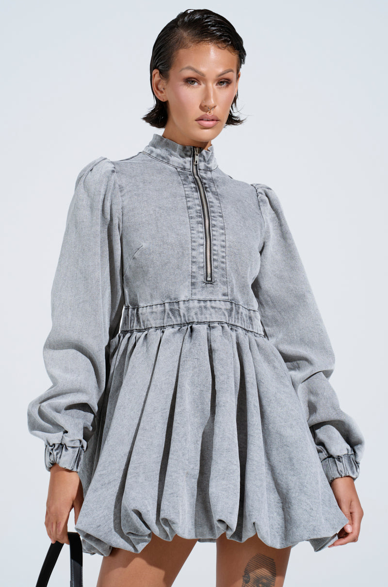 Against a light blue backdrop, a person poses in the PASS THE BUBBLY DENIM MINI DRESS IN LIGHT GREY. The puffy mini dress features long sleeves, a high zippered neck, and a subtle bubble hem for extra flair. A black strap is held by their side.