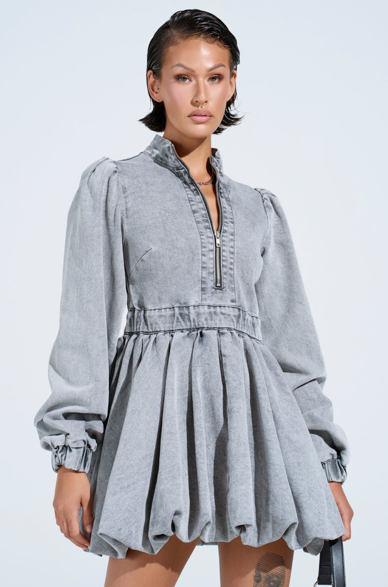 Against a light backdrop, a person stands confidently in the PASS THE BUBBLY denim mini dress in light grey, featuring a balloon-style with bubble hem and front zipper. They showcase short styled hair and minimal accessories.