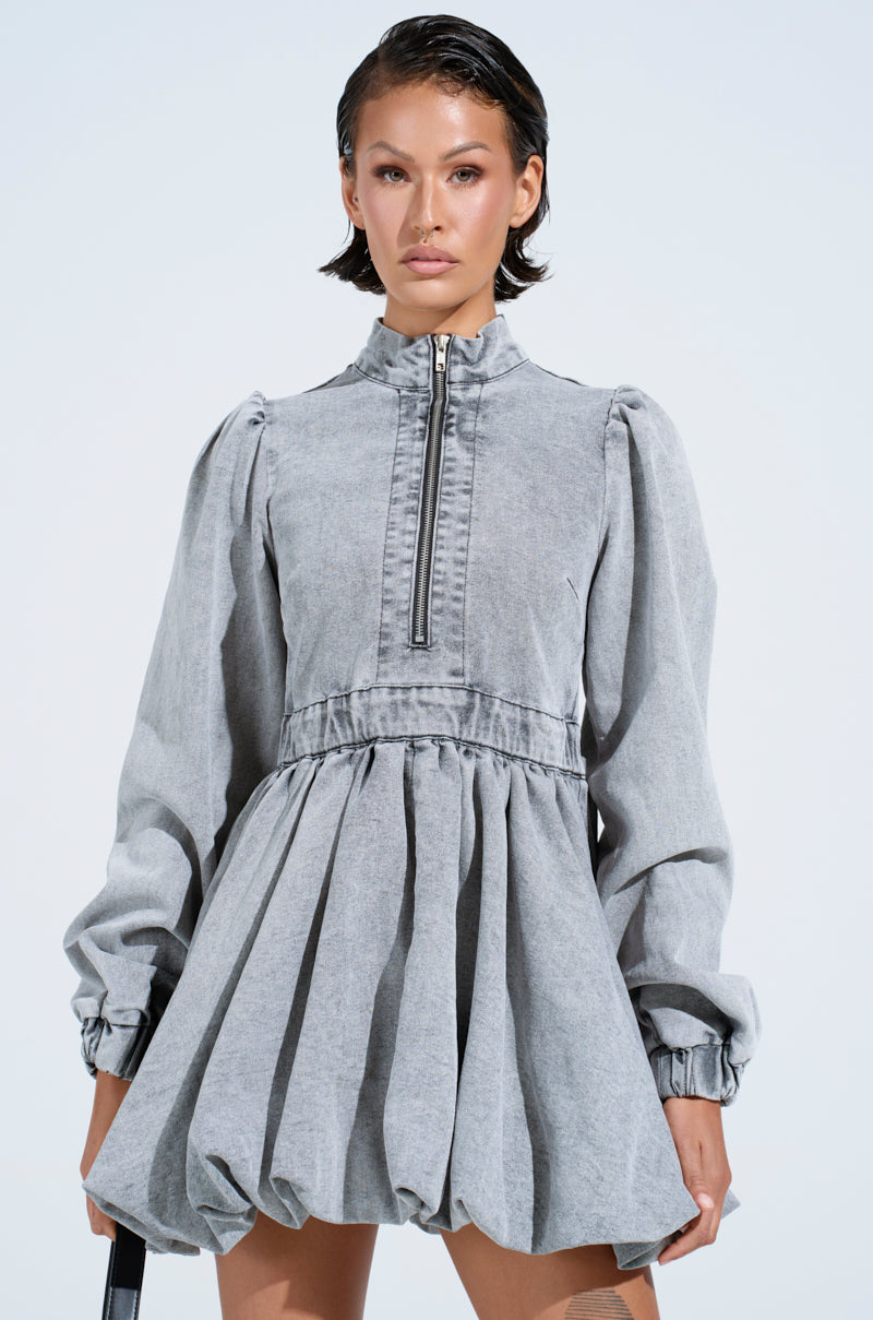 Against a light backdrop, someone confidently wears the PASS THE BUBBLY Denim Mini Dress in light grey, featuring long sleeves, a high neckline, zipper detail, voluminous bubble hem skirt, and gathered waist. They sport short hair and hold a black accessory with flair.