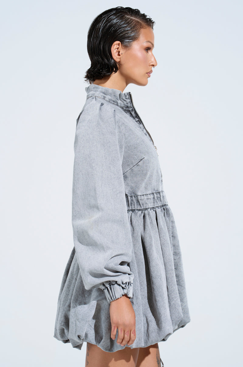Side profile of a person in the PASS THE BUBBLY DENIM MINI DRESS IN LIGHT GREY, featuring a gathered waist and puffed sleeves. The light background highlights the dress’s texture and modern skater silhouette with a playful bubble hem.