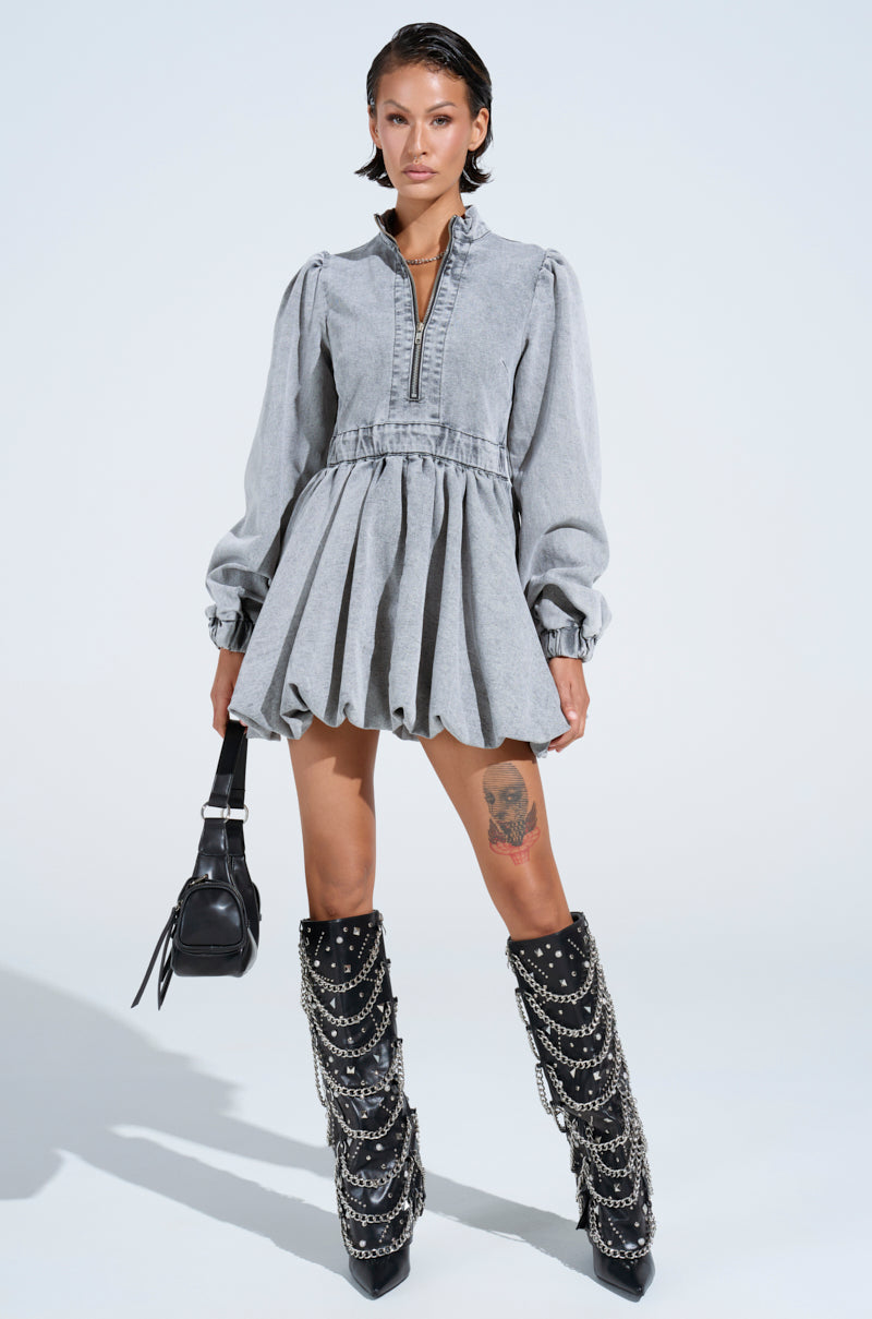 Wearing the "Pass the Bubbly" denim mini dress in light grey with a zippered front, a person holds a black handbag. They sport skater silhouette boots with chains and slicked-back hair that reveals a thigh tattoo against a plain white background, enhancing the edgy look.