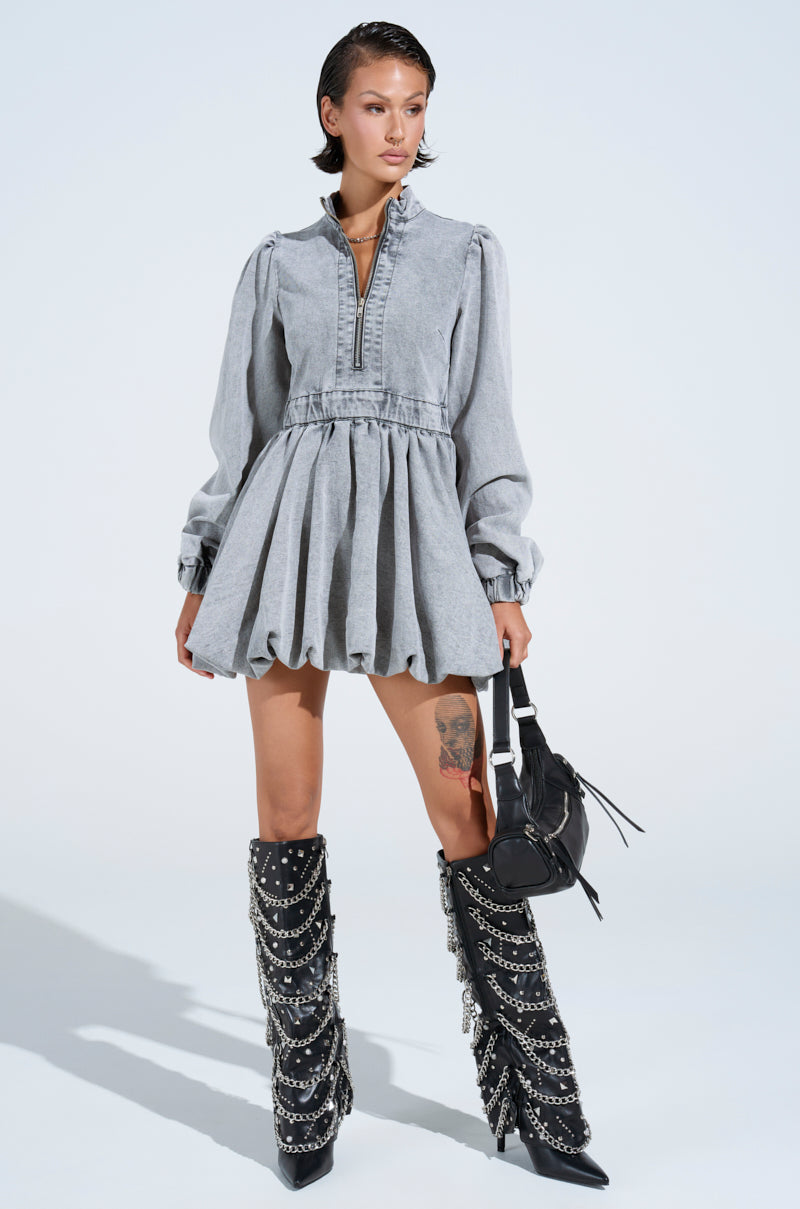 A person confidently wears the PASS THE BUBBLY denim mini dress in light grey with long sleeves and a skater silhouette, knee-high black boots with chains and studs, and holds a black handbag, all set against a plain white background.