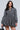A person with long dark hair sports a mini dress similar to the "PASS THE BUBBLY DENIM MINI DRESS," featuring long sleeves and a bubble skirt. They accessorize with a black handbag and a chunky necklace against a light gray background.