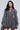 A woman with long dark hair is wearing the "Pass The Bubbly Denim Mini Dress," a gray, long-sleeved skater dress featuring a zippered neckline and a gathered skirt. She accessorizes with a large black stone necklace and carries a black handbag against a plain, light-colored background.