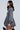 Side view of a person with long dark hair wearing the "Pass The Bubbly" denim mini dress featuring a skater-style silhouette with pleats, standing against a plain, light background.