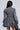 A woman is seen from the back wearing the PASS THE BUBBLY DENIM MINI DRESS, featuring long sleeves and a bubble skirt, while holding a black handbag. Her long dark hair cascades down as she stands against a simple light-colored backdrop.