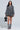 A person stands wearing the PASS THE BUBBLY DENIM MINI DRESS, featuring puffed sleeves and a bubble skirt. They hold a black handbag shaped like a dog and wear matching gray boots. The neutral light gray background perfectly complements the vibe of this chic skater dress ensemble.