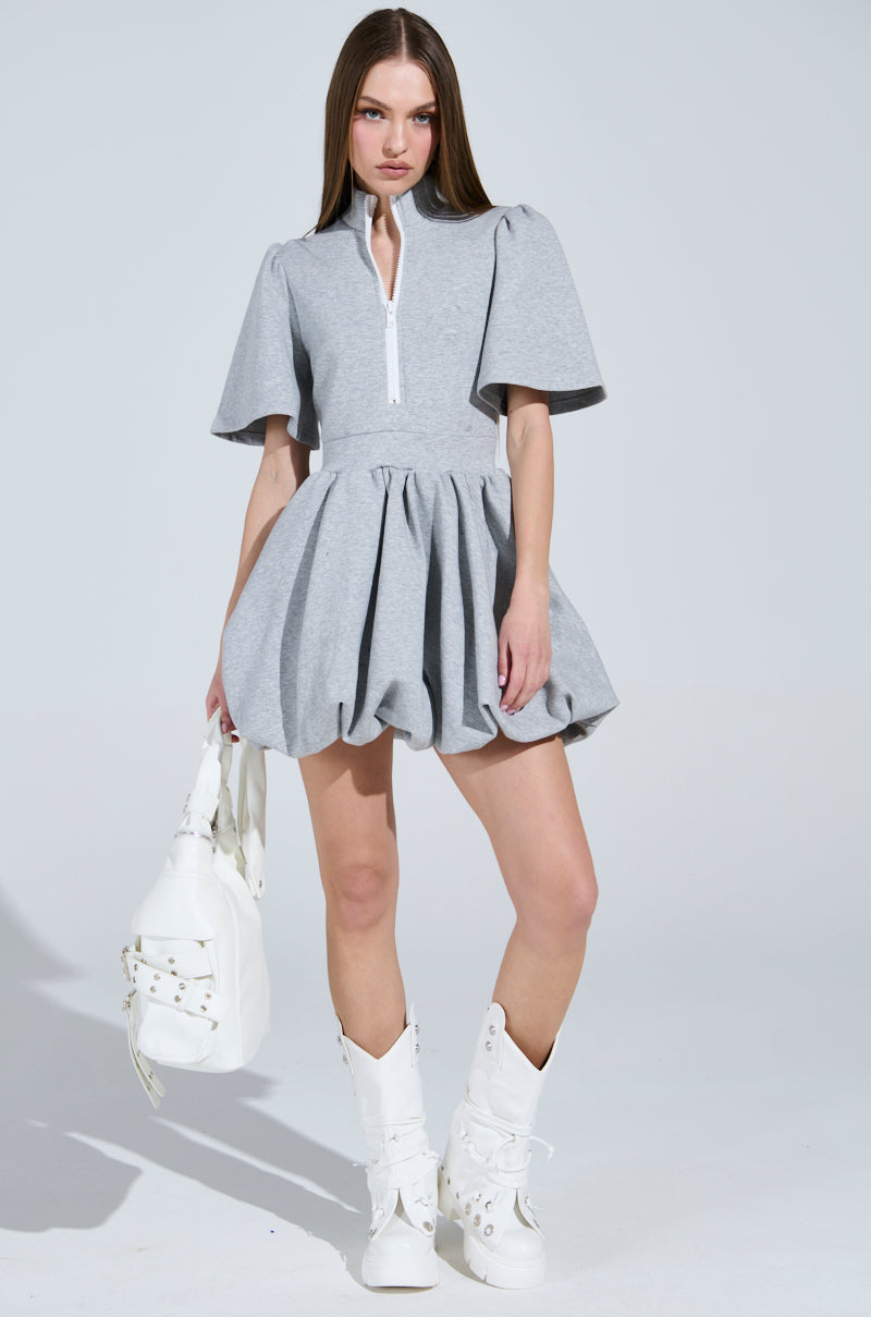 A person confidently poses in the NAME OF THE GAME SHORT SLEEVE ZIPPERED MINI DRESS with puffy sleeves, holding a white bag and wearing tall white boots against a light-colored background.