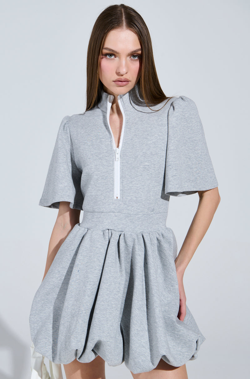 A person poses confidently in the "NAME OF THE GAME" grey mini dress, featuring a zip-up front and puffed sleeves. The pleated, voluminous skirt enhances the flair, while the plain background keeps focus on this stylish ensemble.