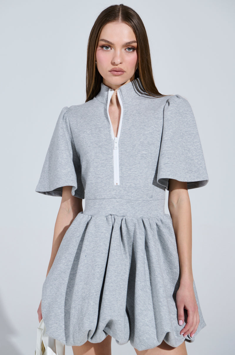 A woman stands against a plain background wearing the NAME OF THE GAME SHORT SLEEVE ZIPPERED MINI DRESS in light gray, featuring a zippered high collar, cinched waist, and voluminous skirt. She holds a white object in her left hand, her long straight hair cascading down as she maintains a neutral expression.
