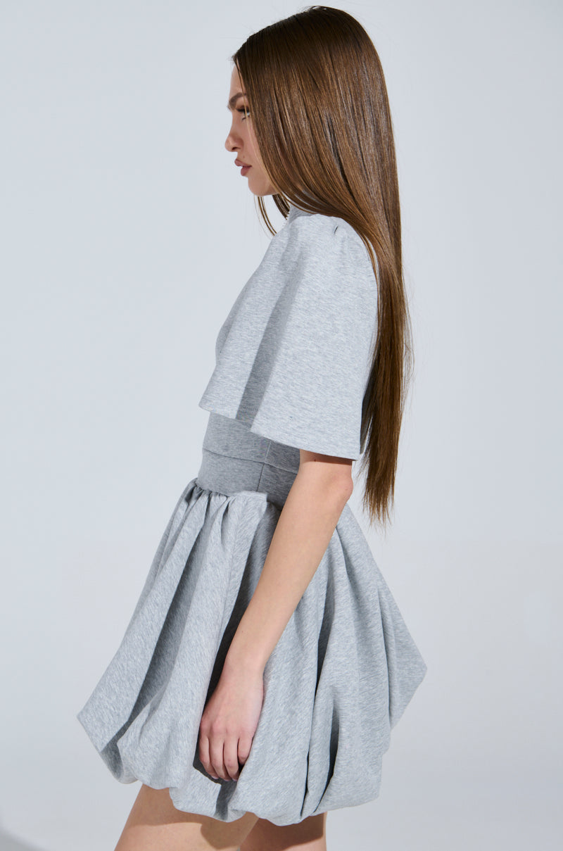 A person with long, straight brown hair is wearing the "Name of the Game" short sleeve zippered mini dress in gray, featuring a chic flared skirt. They are standing sideways against a plain background.