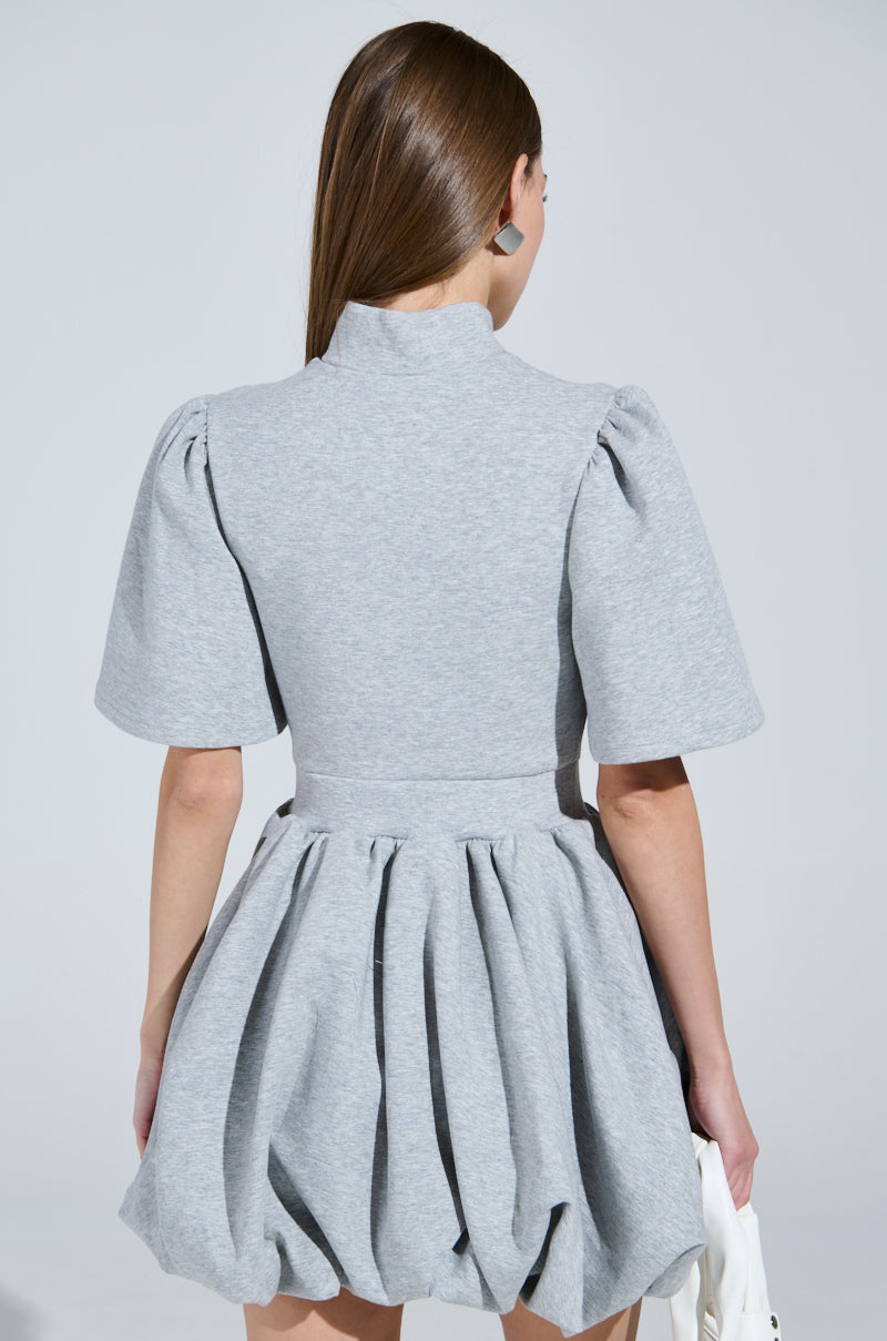 A person with long brown hair models the stylish NAME OF THE GAME SHORT SLEEVE ZIPPERED MINI DRESS in light gray, featuring a chic puffy silhouette and zip-front design. Posed against a plain background, they carry a white handbag, showcasing the dress from the back.