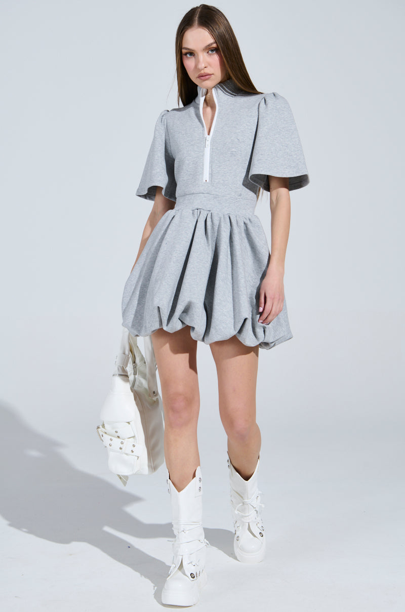 A woman in the "Name of the Game" short sleeve zippered mini dress with puffy sleeves holds a white bag. She completes her chic look with knee-high white boots, set against a plain, light-colored background.