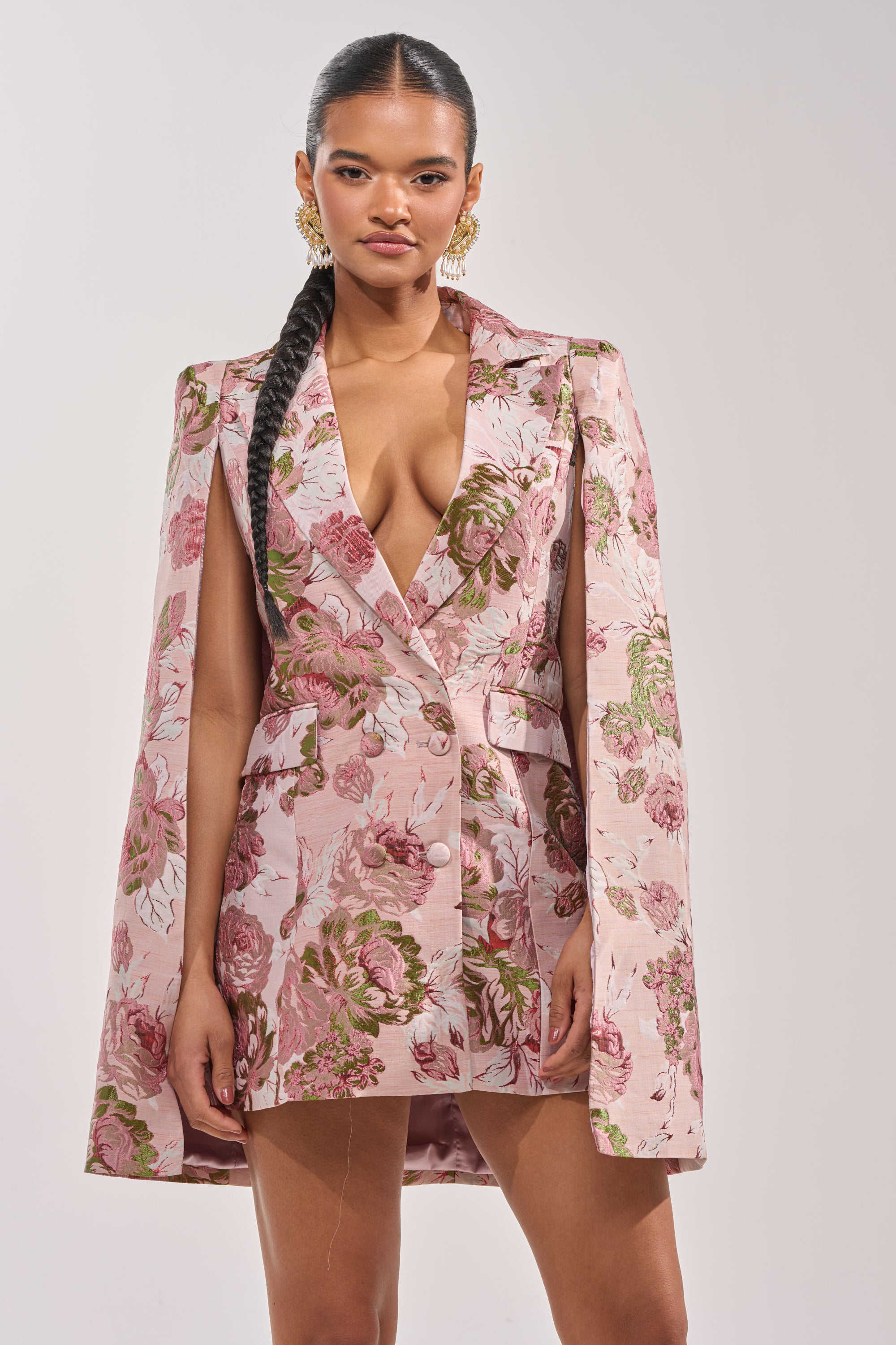 A woman with a long braid wears the IMANI Brocade Cape Sleeve Blazer Dress in Pink, featuring a deep V-neckline and gold statement earrings, posed against a plain light background. 0