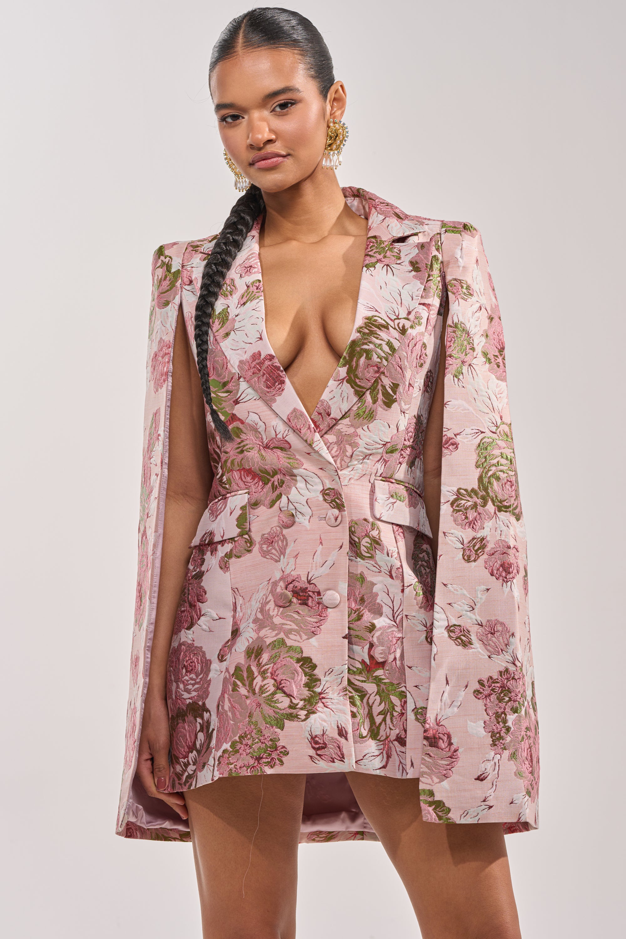 A woman with a long braid stands confidently against a plain light background, wearing the IMANI Brocade Cape Sleeve Blazer Dress in Pink, featuring floral brocade fabric, cape sleeves, and a deep V-neckline. 1