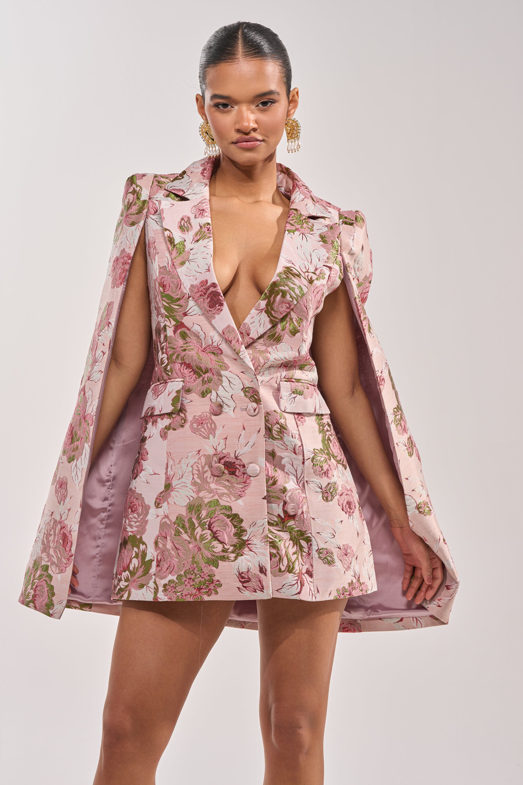 A woman models the IMANI Brocade Cape Sleeve Blazer Dress in pink—a short, sleeveless mini dress with wide lapels and a cape silhouette. She stands confidently, wearing gold earrings against a plain, light background. 2