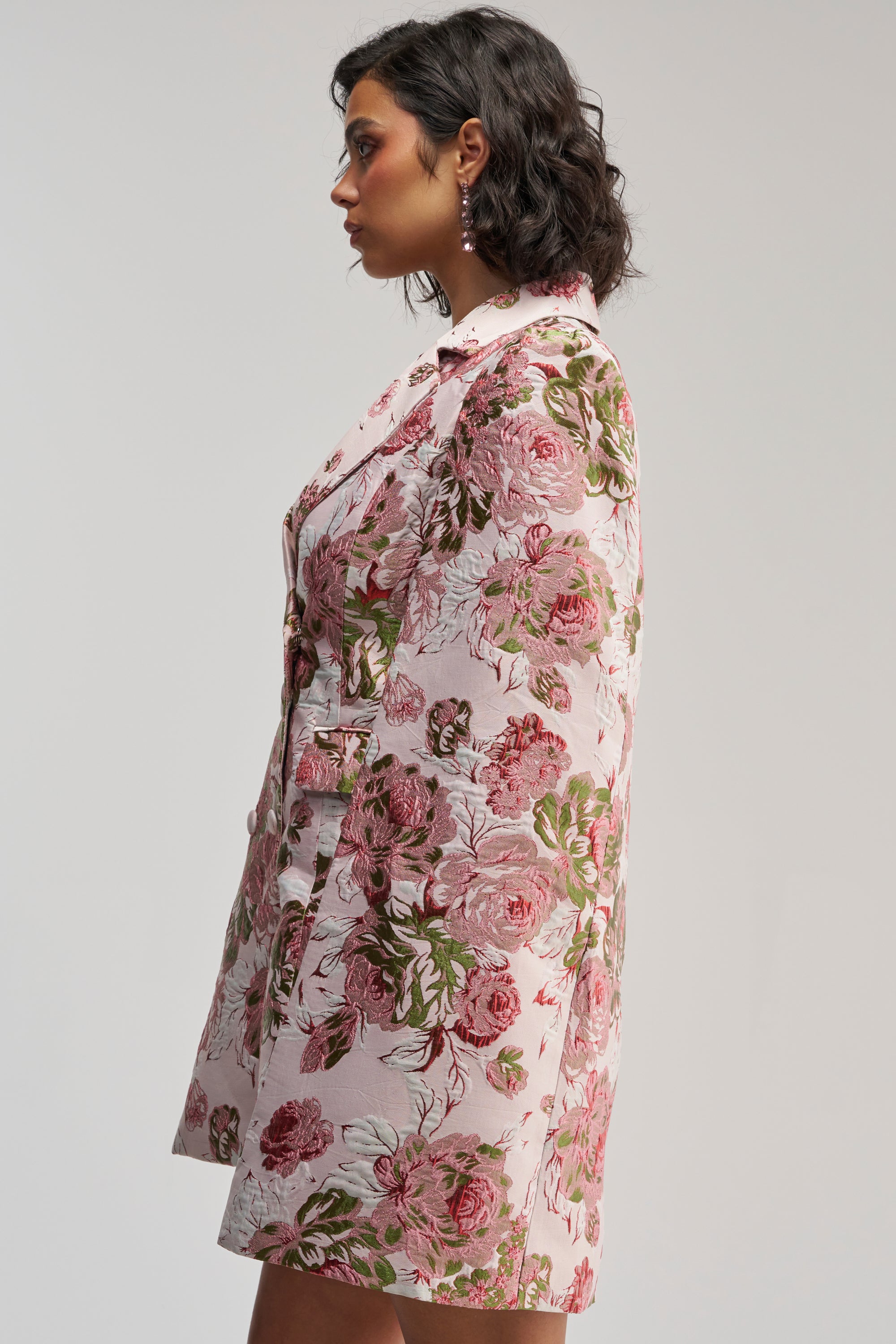 A woman with short dark hair in profile wears the IMANI Brocade Cape Sleeve Blazer Dress in pink, featuring a delicate floral rose pattern, standing against a plain light background.