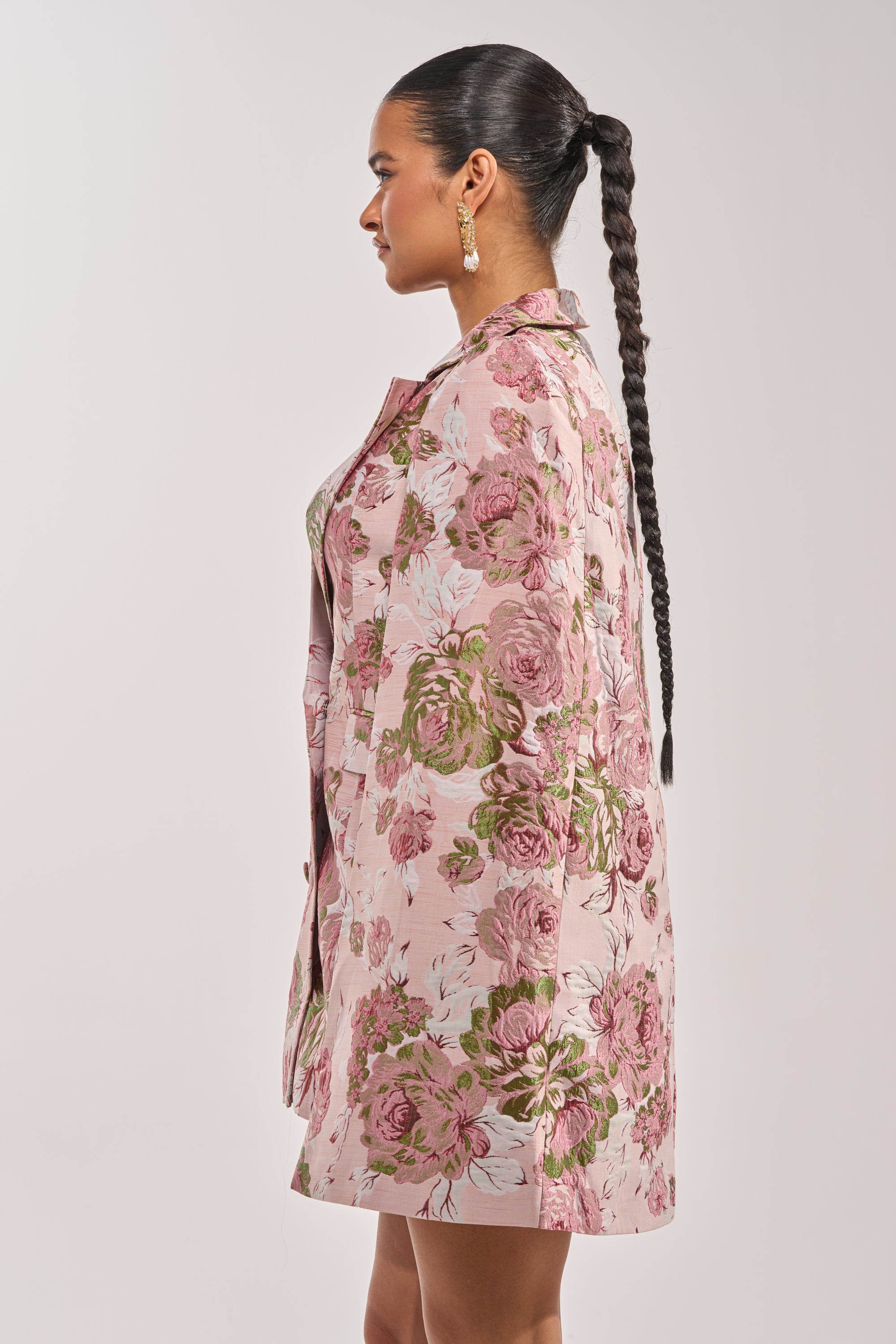 A woman with long braided hair and gold earrings stands in profile against a plain light background, wearing the IMANI Brocade Cape Sleeve Blazer Dress in Pink. 3