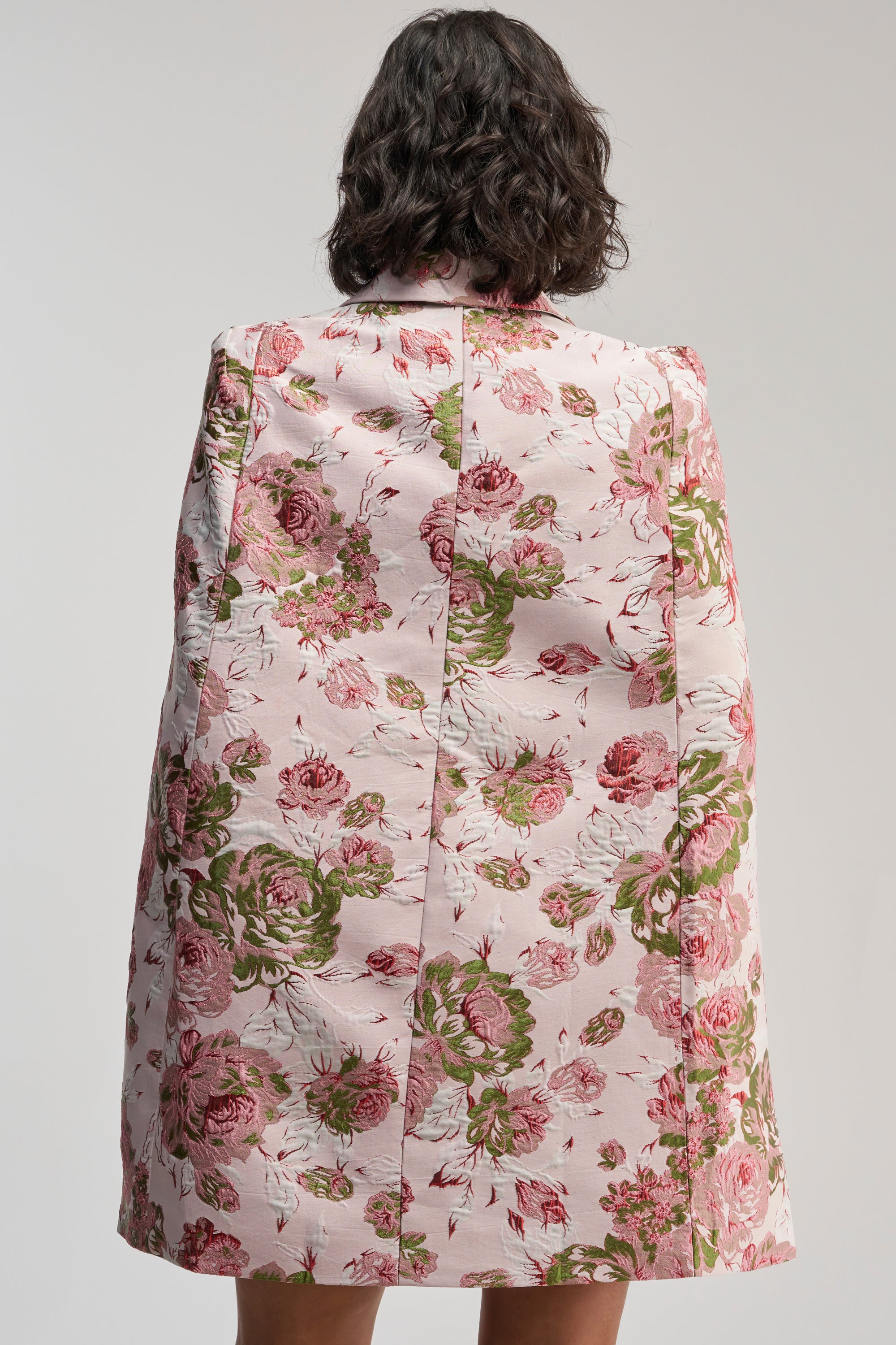 A person with dark wavy hair, seen from behind, wears the IMANI Brocade Cape Sleeve Blazer Dress in Pink, adorned with a light pink floral pattern featuring shades of green and red flowers against a plain backdrop.
