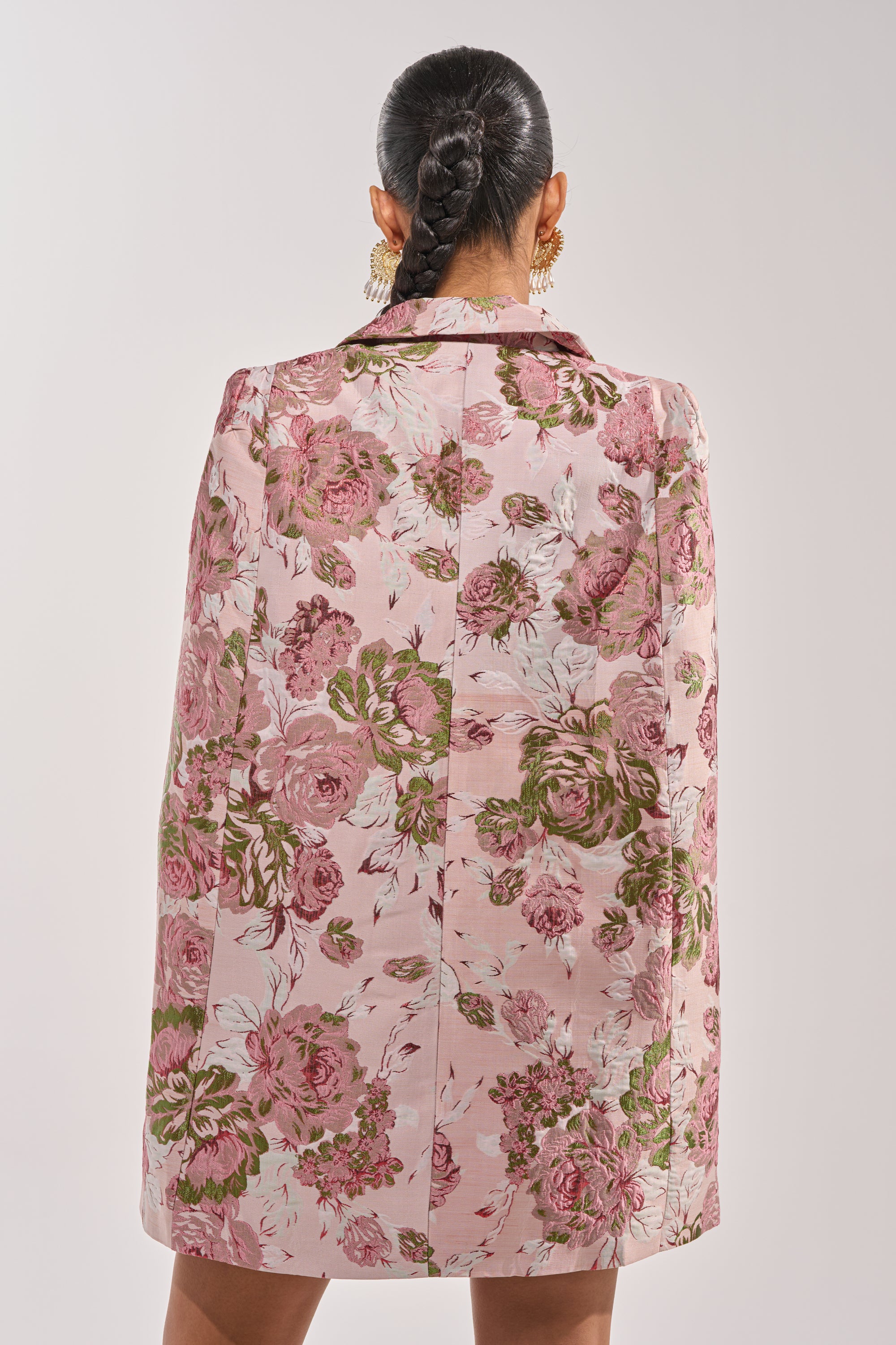 A woman with dark braided hair, seen from behind, wears the IMANI BROCADE CAPE SLEEVE BLAZER DRESS IN PINK with gold statement earrings. 4