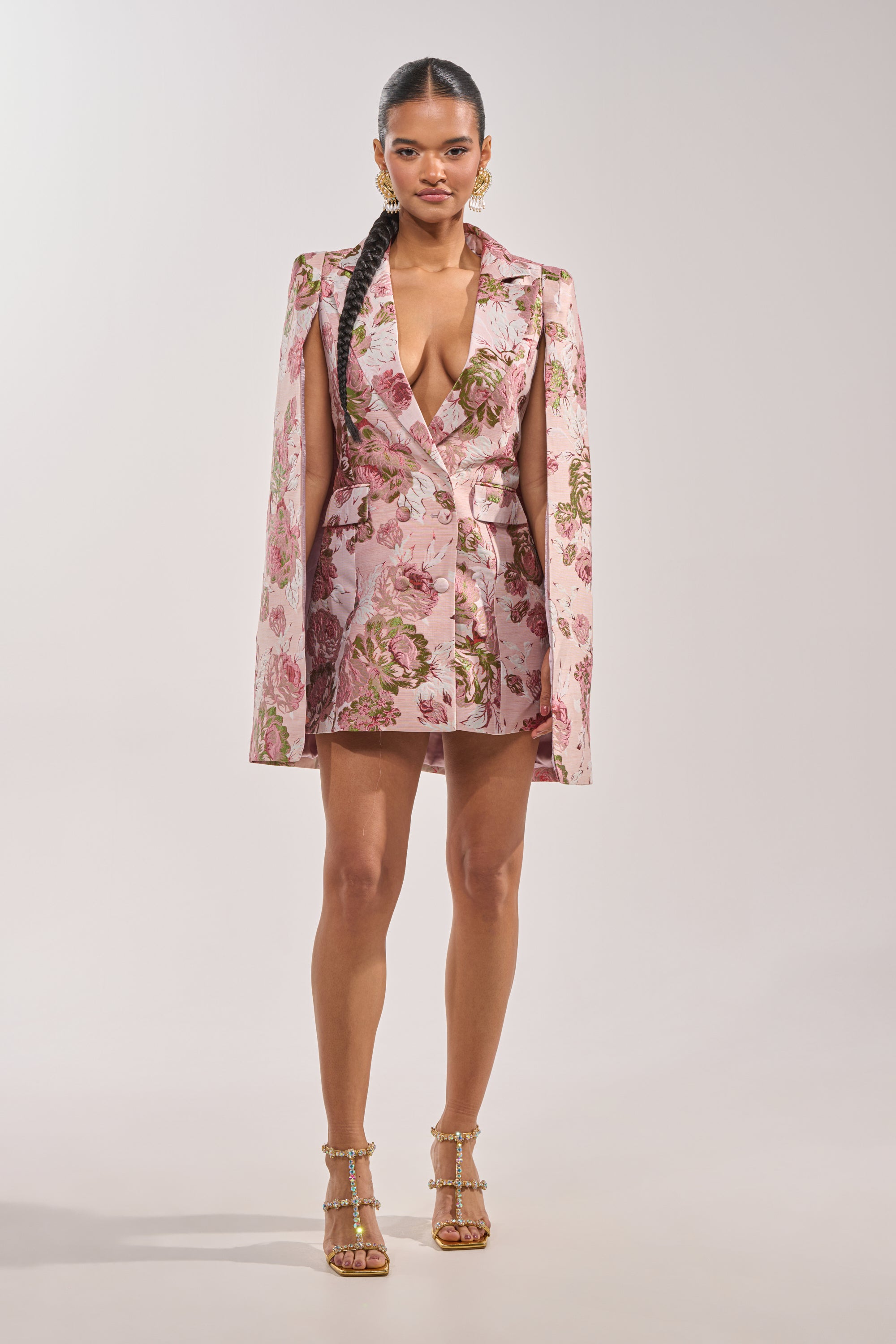 A woman faces forward in the IMANI Brocade Cape Sleeve Blazer Dress in Pink, featuring structured shoulders and cape sleeves. She wears long braided hair, gold earrings, and strappy high-heeled sandals. 5