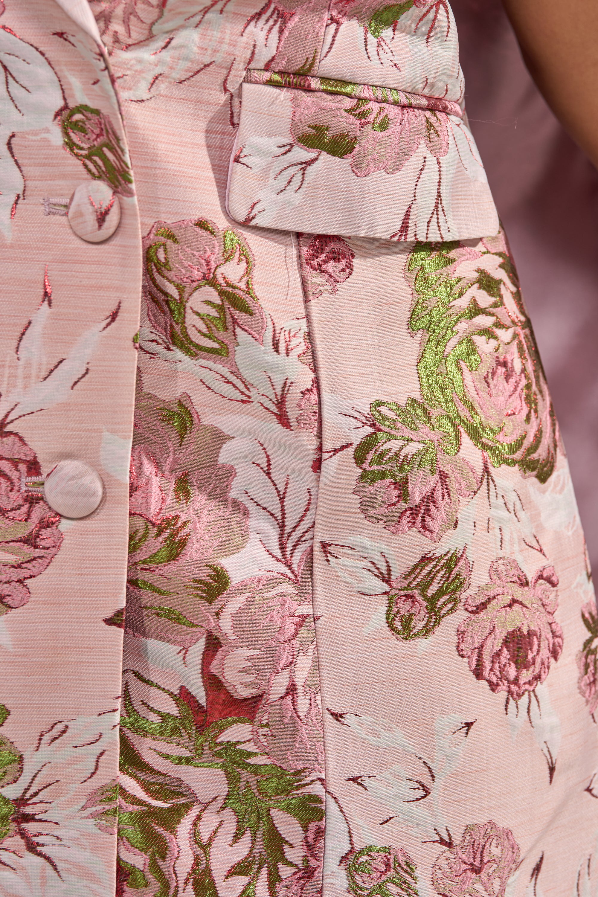 Close-up of the IMANI BROCADE CAPE SLEEVE BLAZER DRESS IN PINK, featuring shiny green and pink embroidered flowers on a pink floral-patterned fabric, with buttons and a flap pocket visible. 7